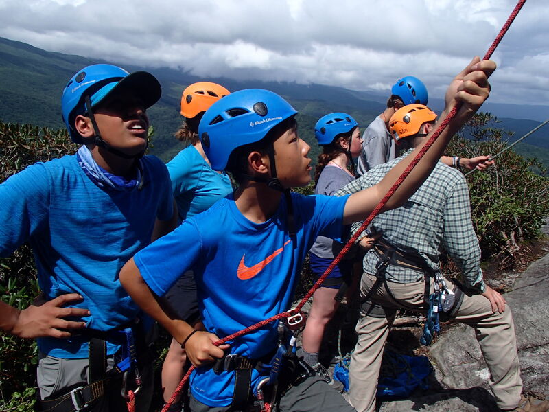 A group of young people are standing on a rocky outcrop, wearing helmets and harnesses, and holding onto a rope. They appear to be engaged in some kind of outdoor adventure activity, possibly rock climbing or rappelling. The background features a scenic view of mountains and trees, suggesting a natural environment.